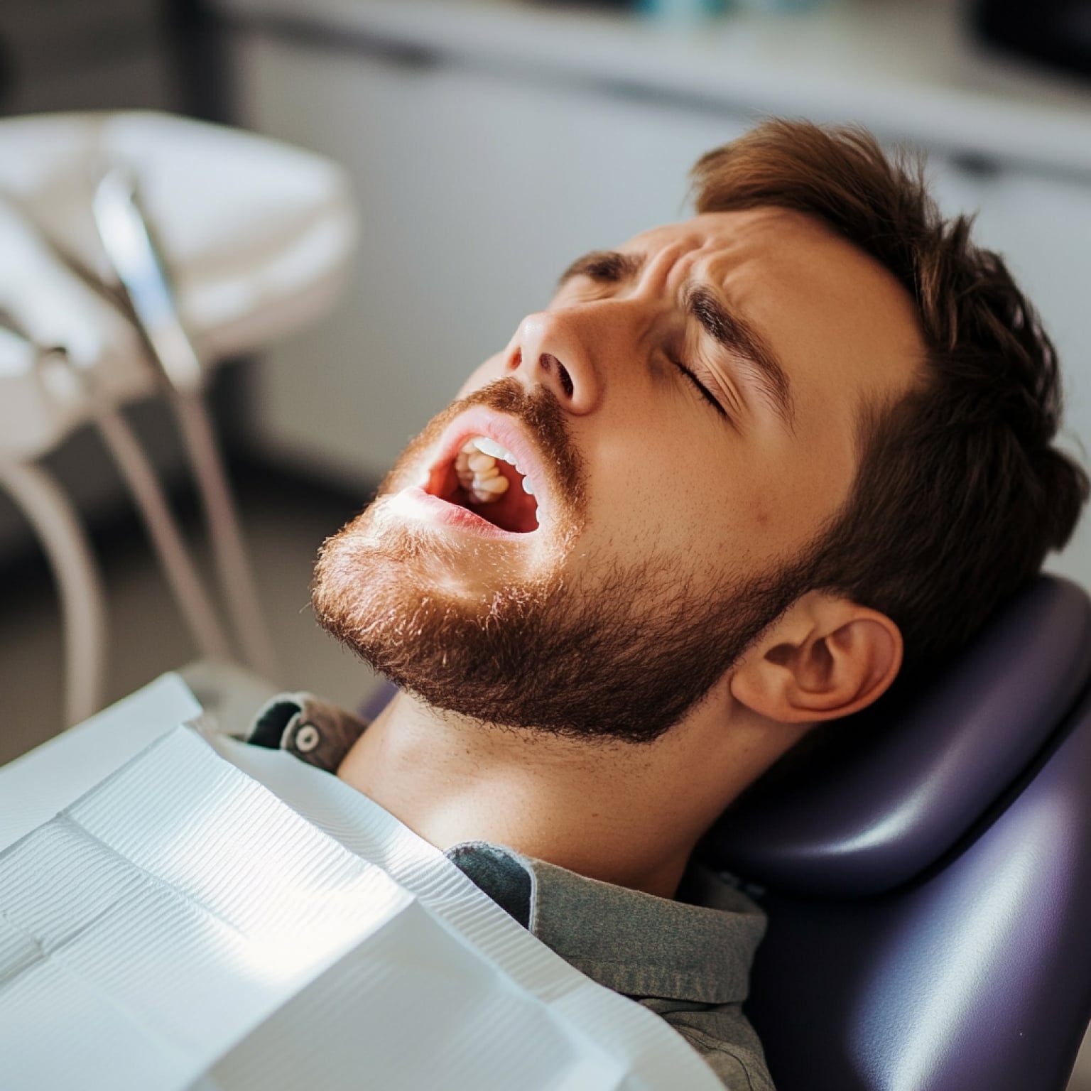 A person reclines in a dental chair with mouth open, wearing a protective bib, in a brightly lit clinical setting.